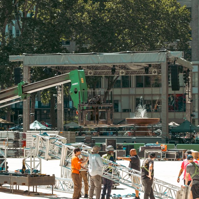Workers assembling an outdoor stage in a New York City plaza, with visible truss and equipment.